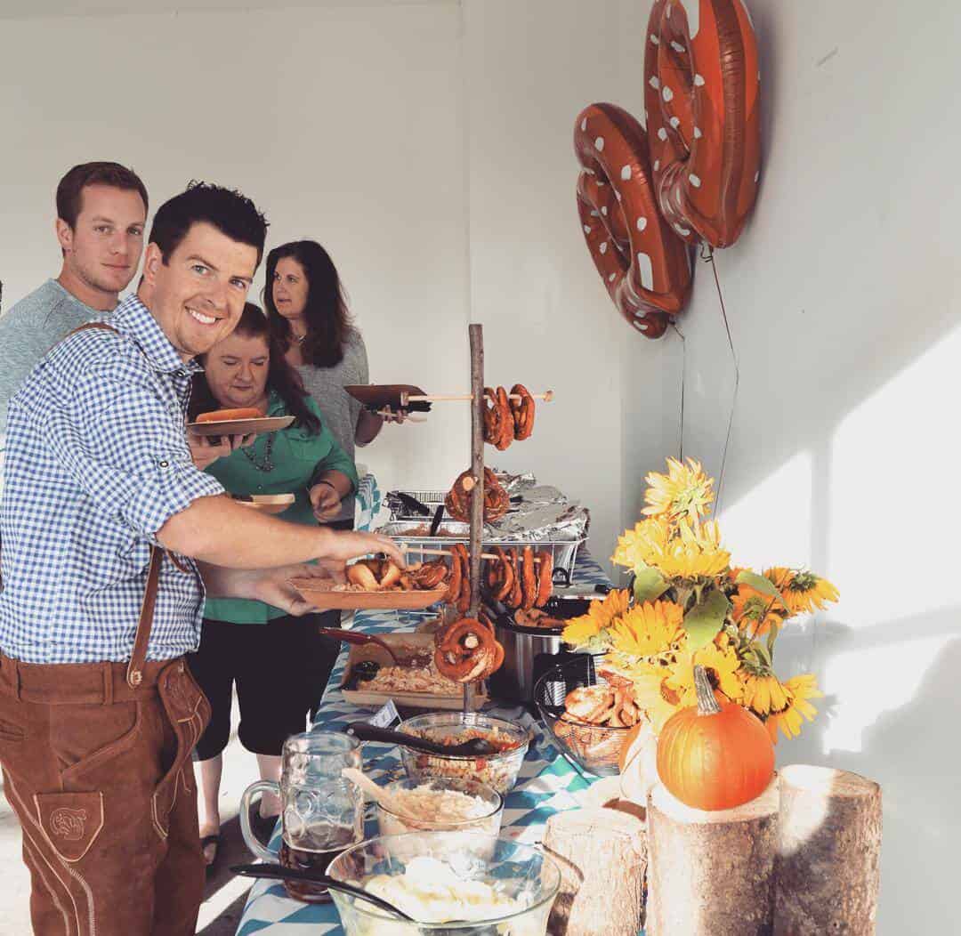 man dressed in lederhosen standing next to Oktoberfest party buffet