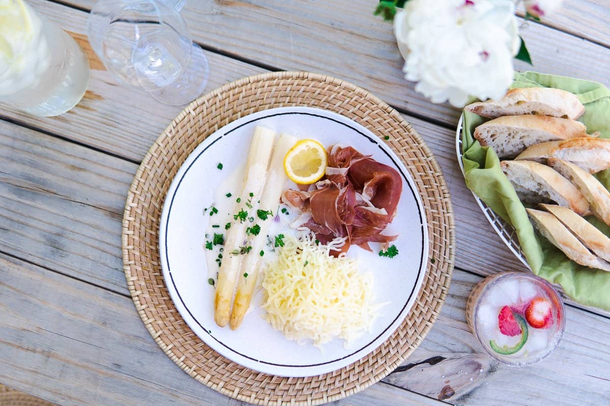 a plate with white asparagus, ham and potatoes, a cocktail and a bread basket