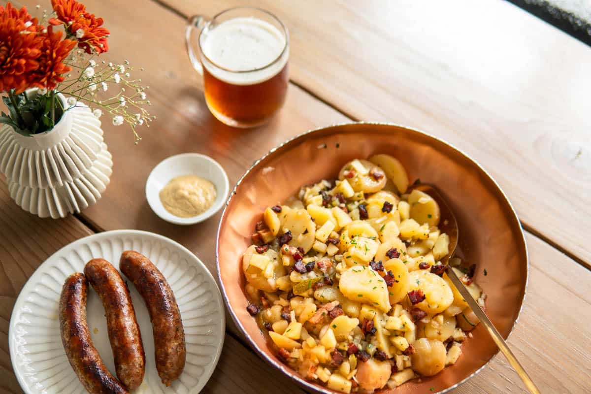 bowl of potato salad next to plate with grilled sausages and a glass of beer