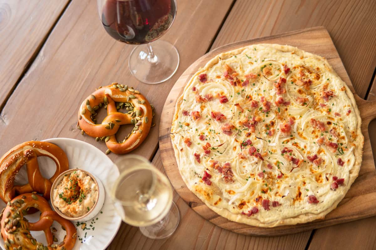 Flammkuchen on a serving board, pretzel on a table next to it and a glass of red wine
