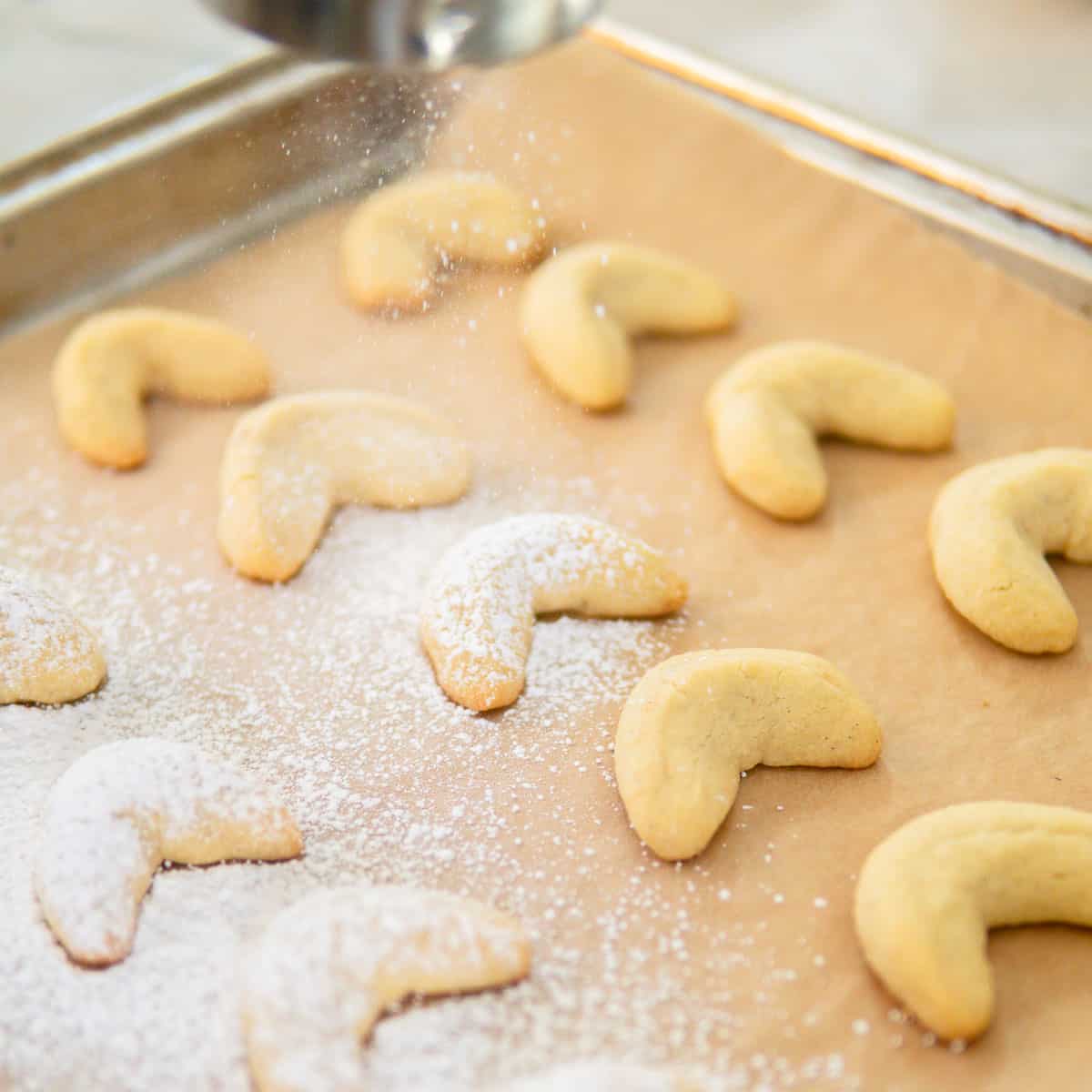 Vanillekipferl on a baking sheet getting a powdered sugar dusting