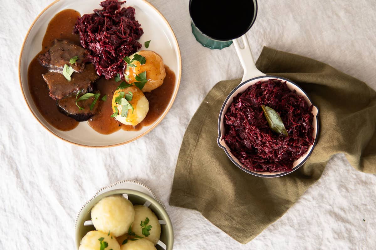 German red cabbage in a small skillet next to a plate with sliced beef roast, potato dumplings and red cabbage.