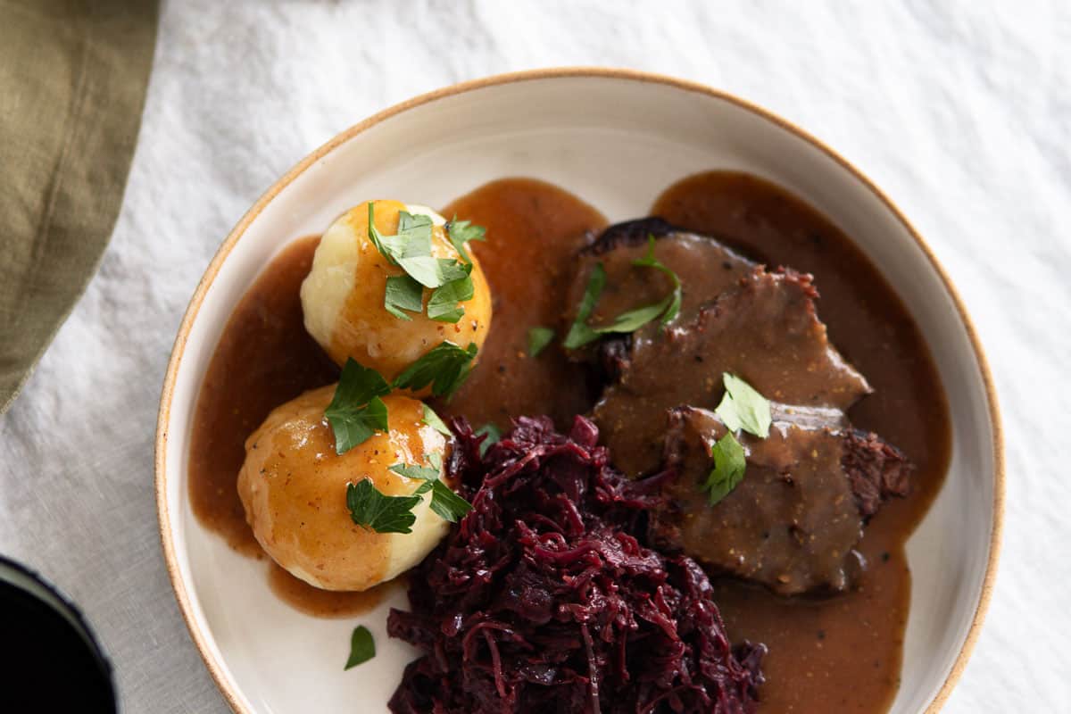 Closeup of a dinner plate with red cabbage, beef roast, sauce and potato dumplings.