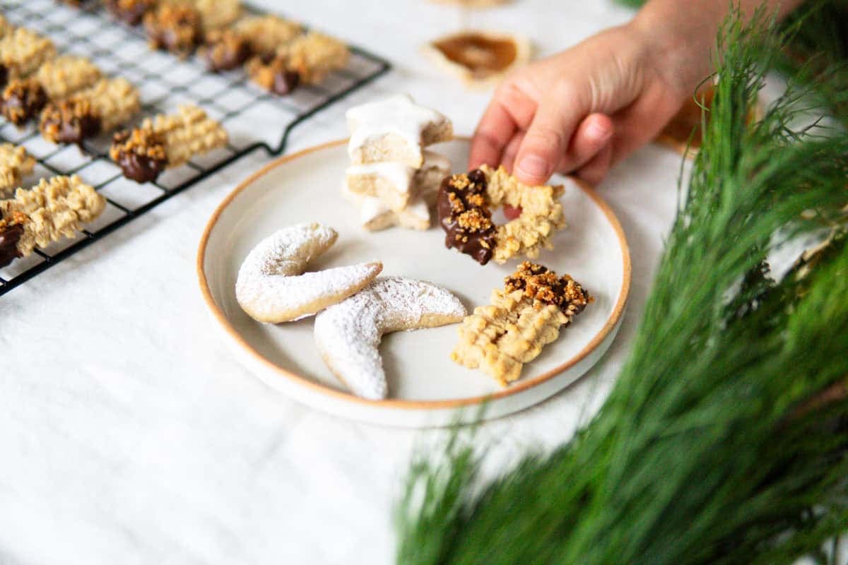 A child holding a ring-shaped Spritz cookie dipped in chocolate next to a plate of German Christmas cookies.