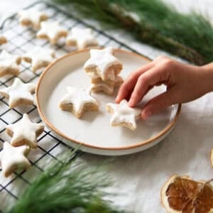 A child holding a star shaped cinnamon cookie called Zimtsterne next to a cooling rack full of Zimtsterne.
