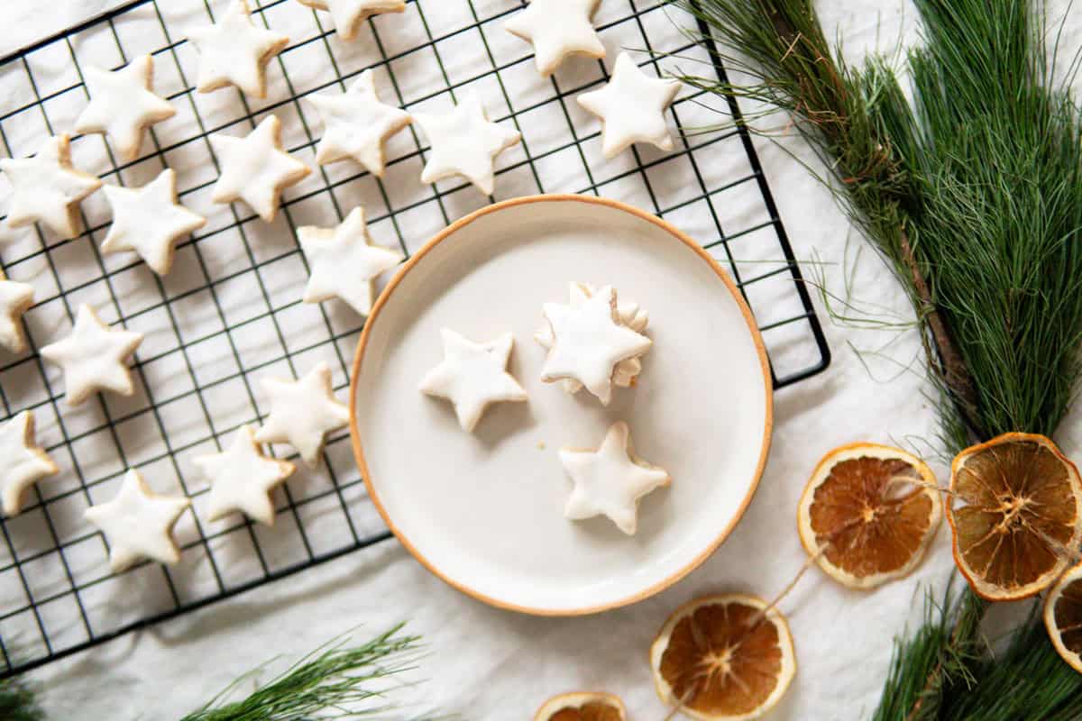 Star shaped cinnamon cookies called Zimtsterne on a small plate next to a cooling rack full of Zimtsterne.