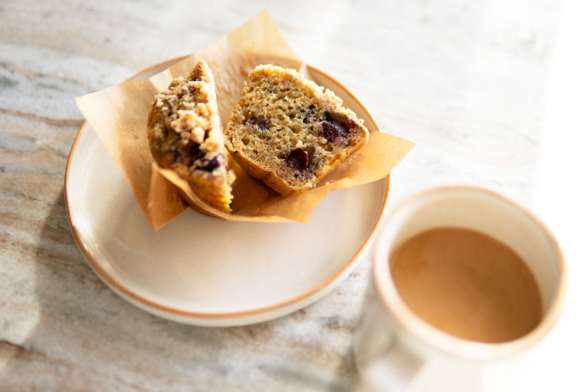 Banana muffin cut in half on a small plate next to a cup of coffee.