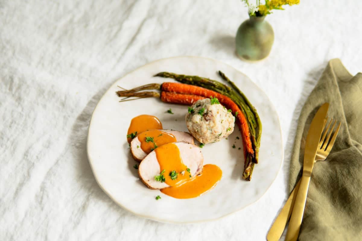 Sliced ​​pork loin on a plate with sauce, a bread dumpling and roasted veggies.