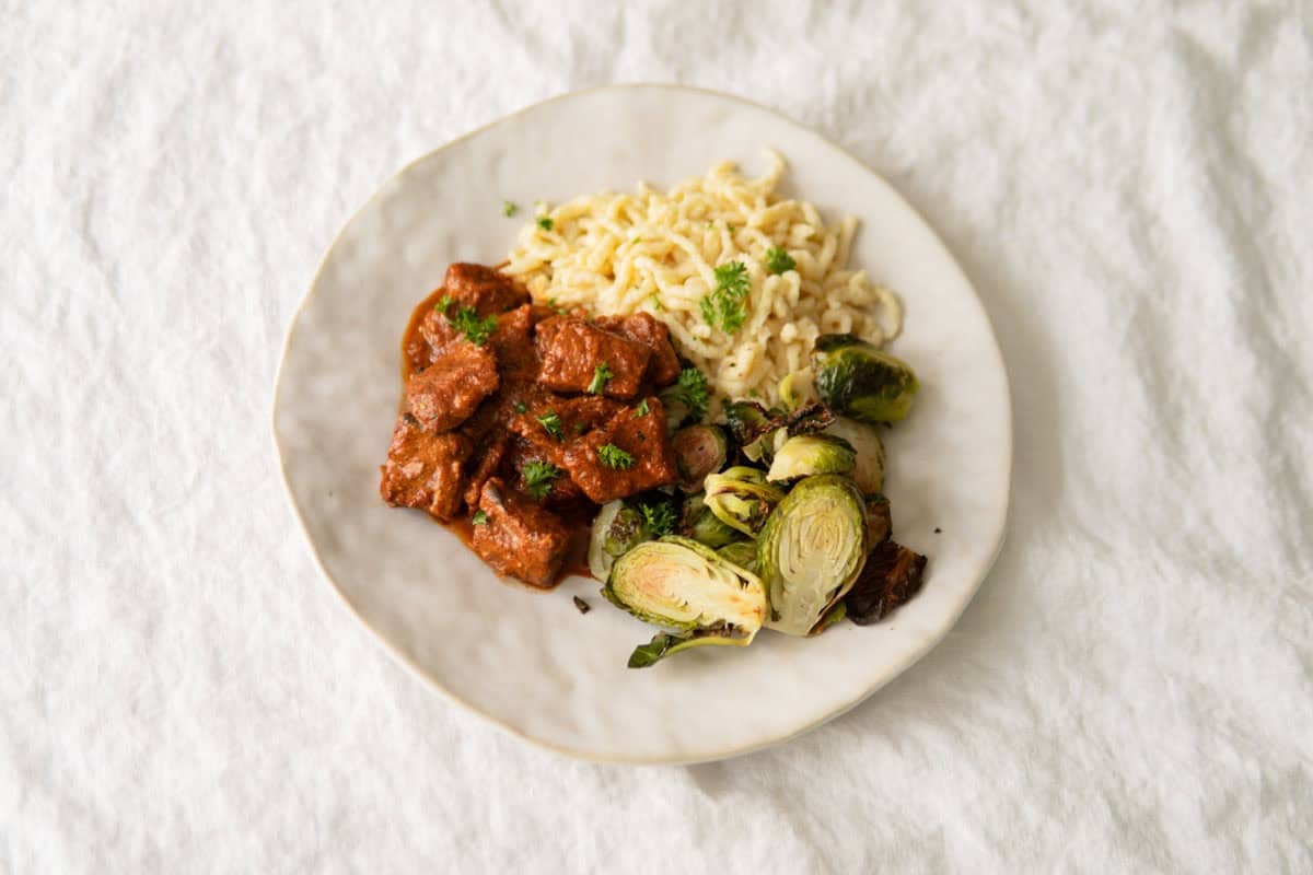 German pork goulash on a plate with spätzle noodles and brussel sprouts.